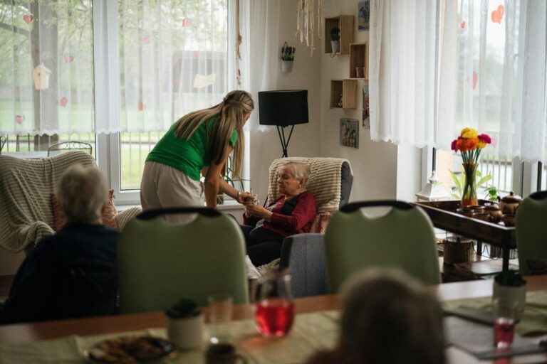 Une soignante se penche vers une résidente assise dans la salle commune d'un établissement