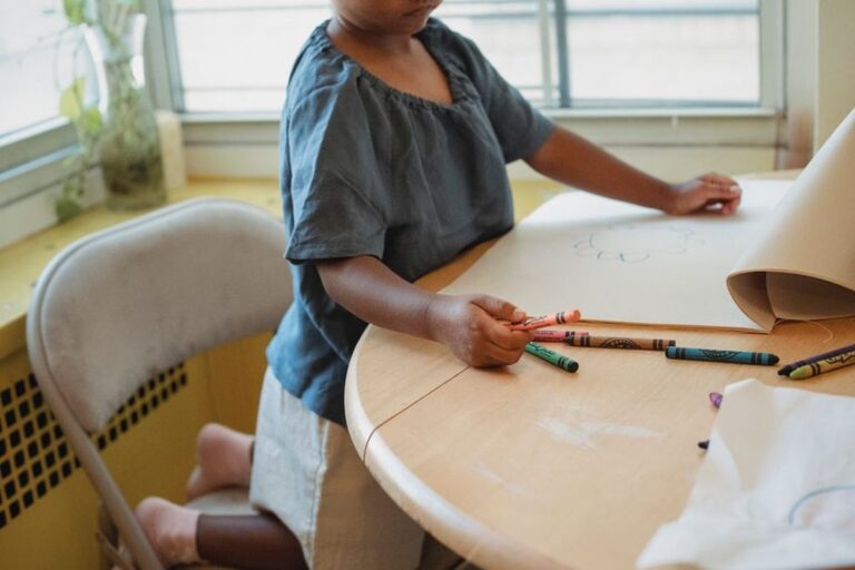 Un enfant debout à une table dessine aux crayons de couleur près d'une fenêtre